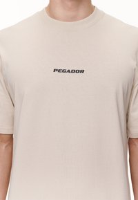Beige lyhythihainen pyöreäkauluksinen t-paita, jossa on mustalla painettuna sana "PEGADOR" keskellä rintaa.