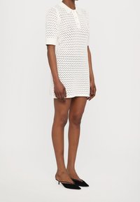 Robe blanche à manches courtes avec un col et un motif perforé. Associée à des chaussures noires à enfiler.