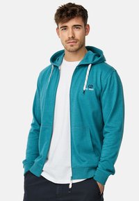 Veste à capuche zippée bleu canard en tissu doux avec une poche avant, cordons de serrage blancs et détail de logo sur la poitrine. Portée sur un t-shirt blanc.