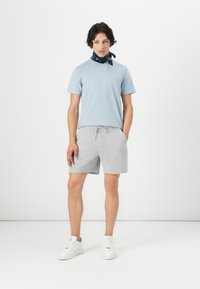 T-shirt bleu clair à manches courtes, shorts rayés gris et blancs, baskets blanches, et un bandana à motifs sombres autour du cou.