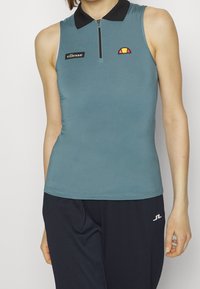 Ärmlös petrolblå polo-topp med svart krage, dragkedja framtill och broderad logotyp. Slät textur med minimala sömnadsdetaljer.