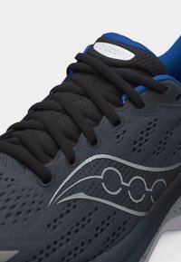 Κλείσιμο μιας μαύρης και μπλε αθλητικής παπούτσας Saucony με μαύρα κορδόνια, ανθεκτικό διχτυωτό επάνω μέρος και ασημένιο λογότυπο στο πλάι.