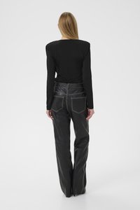 Blond kvinne som står med ryggen mot, iført en svart langermet topp og svarte skinnbukser med hvit søm mot en ensfarget bakgrunn.