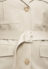 Μπεζ παλτό με μεγάλα κουμπιά, δύο τσέπες με καπάκι και φαρδιά υφασμάτινη ζώνη στερεωμένη με ορθογώνια αγκράφα στη μέση.