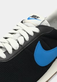 Ténis Nike com parte superior em corduroy preto, swoosh azul, atacadores brancos e língua almofadada; apresenta superfície texturizada e sola de borracha.