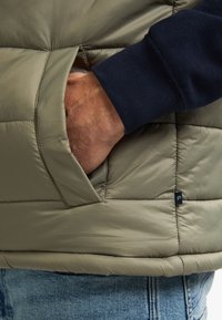 Helle olivgrüne gepolsterte Weste mit Seitentasche, strukturiertem Finish und navyblauem Bund. Verfügt über ein kleines Logotag nahe dem Saum.