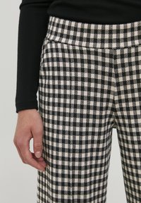 Un pantalon à carreaux noir et crème avec une texture lisse, présentant une taille ajustée et des poches latérales, exposé avec une main posée à proximité.