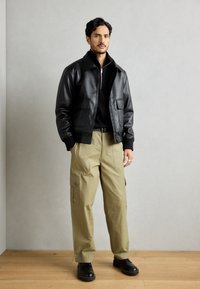 Čierna kožená bomber bunda s dvoma prednými vreckami, nosená cez čierny zipsový sveter, skombinovaná s khaki cargo nohavicami a čiernymi topánkami.