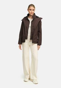 Vrouw staat rechtop in een donkerbruine jas met een ritssluiting aan de voorkant, een beige top, crèmekleurige wijde broek en witte sneakers tegen een effen achtergrond.