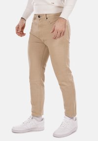 Pantaloni slim-fit khaki in cotone, caratterizzati da cinque tasche, chiusura con bottone e dettagli di cucitura sottili. Indossati con sneakers bianche.
