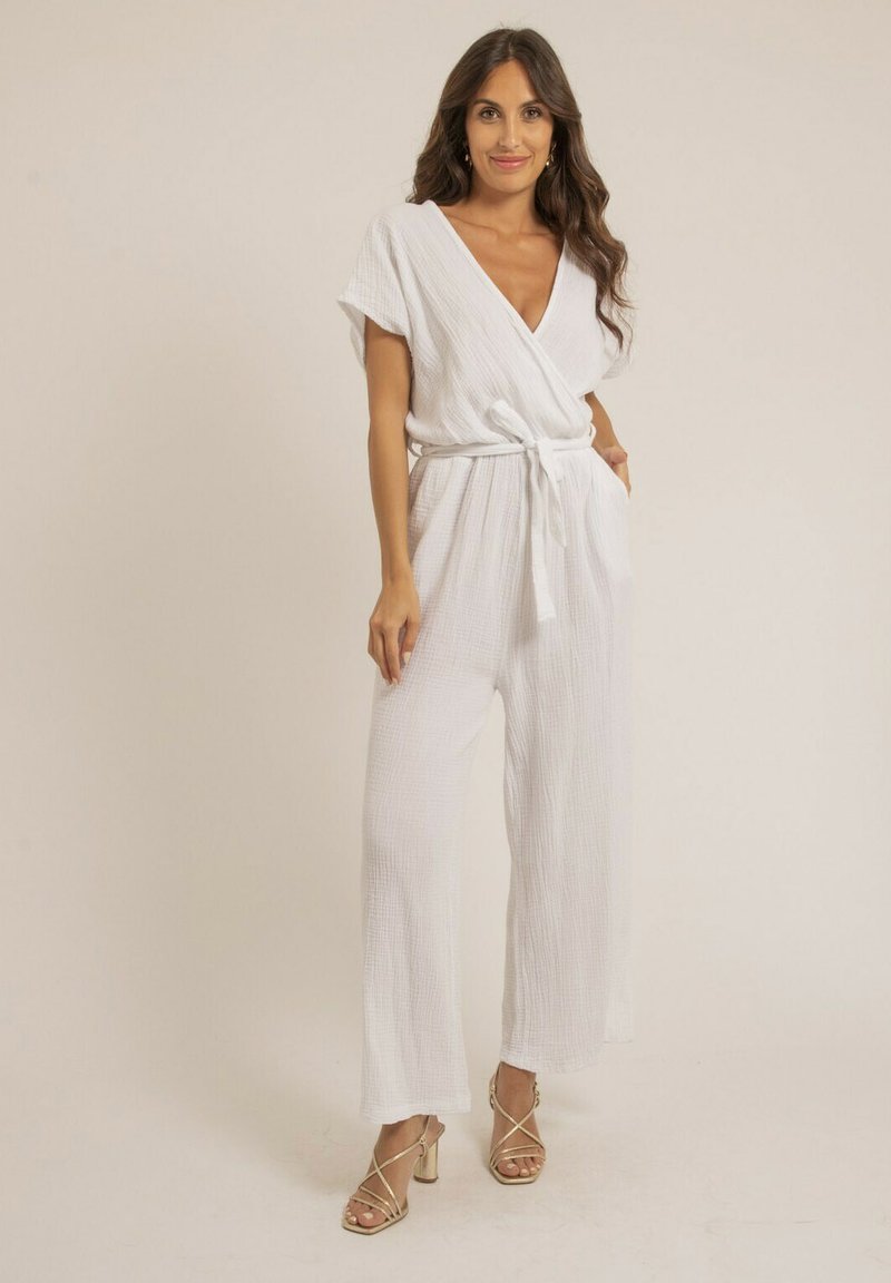 Jumpsuit blanc à manches courtes, en tissu texturé, avec une taille ceinturée et des jambes larges. Associé à des sandales à talons dorées.