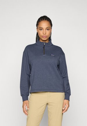 Jonge vrouw met gevlochten haar, gekleed in een marineblauwe fleece met een kwart rits en beige broek, staande tegen een effen lichte achtergrond.