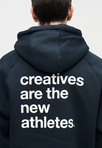 Person facing away wearing a dark hoodie with a white text on the back: "creatives are the new athletes."