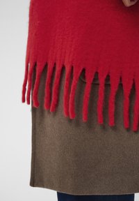 Red wool poncho with fringe detail at the bottom, worn over a brown fabric underneath, partially obscured. Soft texture and layered design.