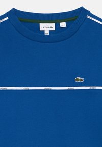 Modrá crewneck košeľa Lacoste s bielym logom pásky cez hrudník a ramená, s malým zeleným logom krokodíla na ľavej strane hrudníka.