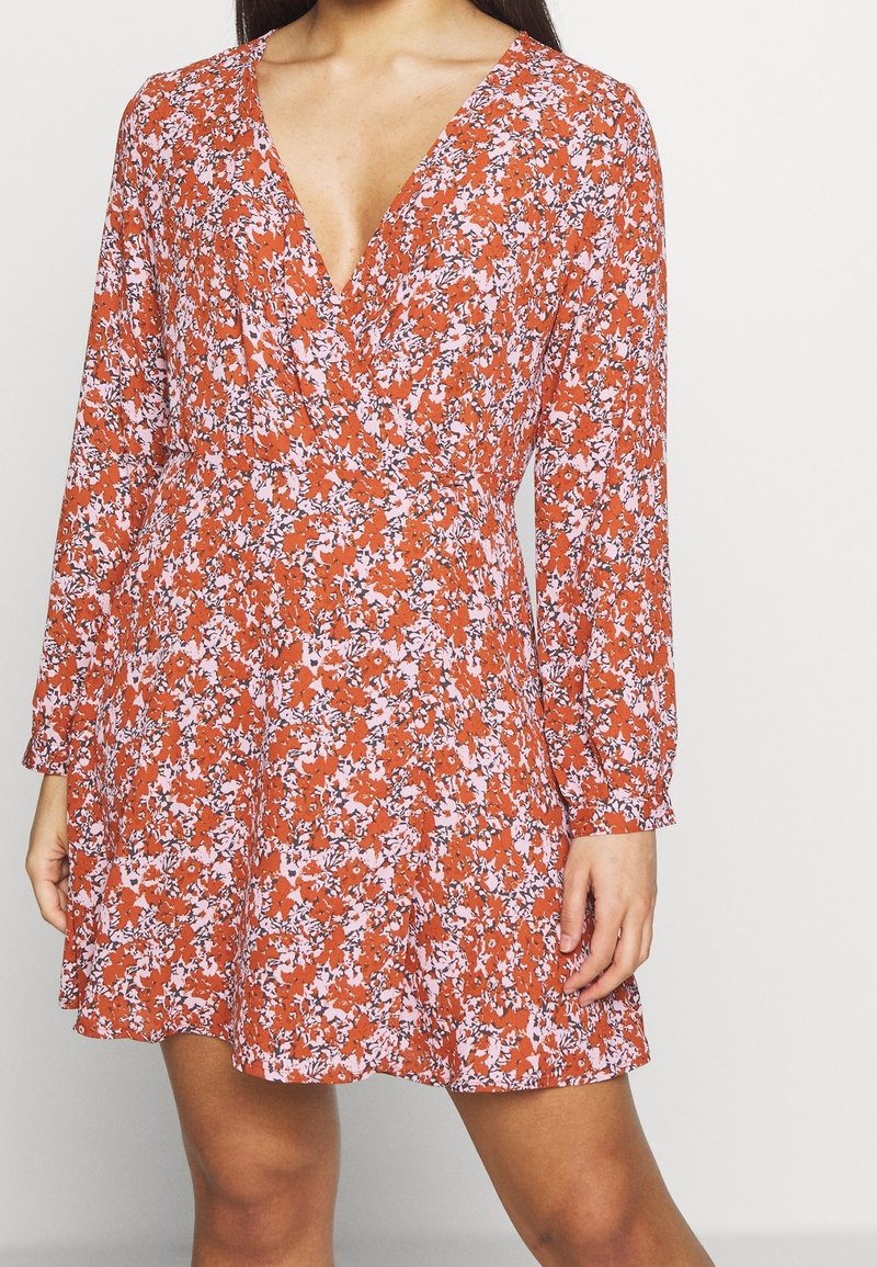 Femme portant une robe courte à manches longues, col en V, avec un motif floral orange et blanc, debout devant un fond uni.