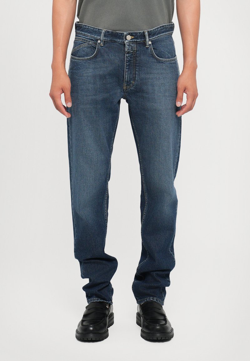 Jeans din denim albastru cu croi drept, cinci buzunare, detaliu subtil de uzură și cusături contrastante. Poartă pantofi negri slip-on.
