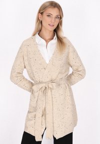 Cardigan a maglia beige con macchie scure, apertura frontale, maniche lunghe e una cintura legata in vita. Texture morbida, forma allungata.