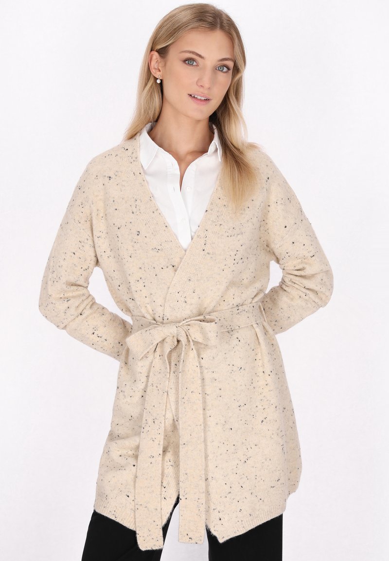Cardigan a maglia beige con macchie scure, apertura frontale, maniche lunghe e una cintura legata in vita. Texture morbida, forma allungata.