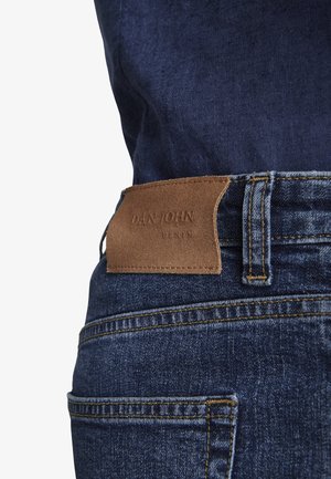 Nahaufnahme einer dunkelblauen Jeansrückentasche mit braunem Leder-Logoaufnäher und sichtbaren Gürtelschlaufen-Nähten.