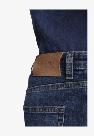 Nahaufnahme einer dunkelblauen Jeansrückentasche mit braunem Leder-Logoaufnäher und sichtbaren Gürtelschlaufen-Nähten.