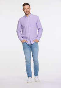 Camisa de botón en color lila claro con un bolsillo en el pecho, combinada con jeans de color azul claro y zapatillas blancas. La tela parece suave y ligeramente texturizada.