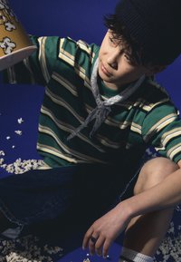 Striped green and yellow shirt, denim shorts, gray bandana, and black beanie. Spilling popcorn on a blue background. Hand with a ring.