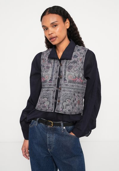 Γκρίζα γιλέκο με περίπλοκα σχέδια, διαθέτοντας κουμπιά μπροστά, δομημένο γιακά και στενή σιλουέτα πάνω από ένα σκούρο πουκάμισο.