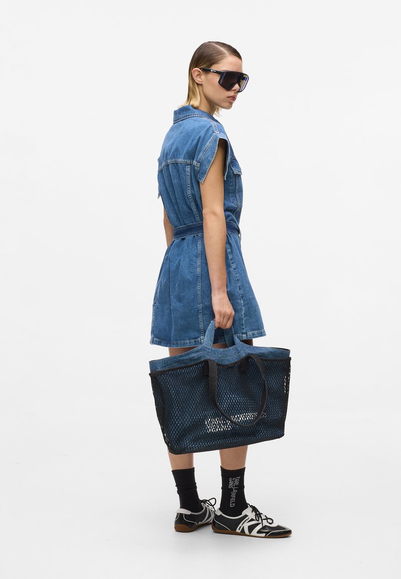 Γυναίκα που φοράει αμάνικο φόρεμα από τζιν και μεγάλα γυαλιά ηλίου, κρατώντας μπλε τσάντα από δίχτυ, μαύρες κάλτσες και μαύρα-άσπρα αθλητικά παπούτσια.