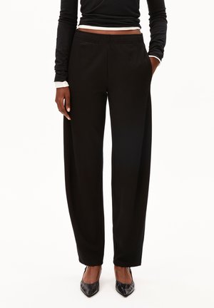 Persoon die zwarte brede broek, zwarte top met lange mouwen en zwarte puntige schoenen draagt, staand met één hand in de zak.