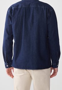 Mörk denimskjorta med långa ärmar, knäppta manschetter och rak passform. Har krage och synlig sömnad, bärs med ljusa byxor.