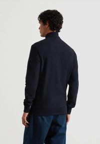 Suéter azul marino con cuello alto y mangas largas, que presenta una tela texturizada. Se ajusta cómodamente a la cintura, combinado con pantalones azules holgados.