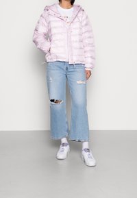 Giacca a vento rosa chiaro con cappuccio, caratterizzata da un design trapuntato e cerniera frontale. Indossata sopra jeans a gamba larga azzurri chiari strappati e scarpe bianche.