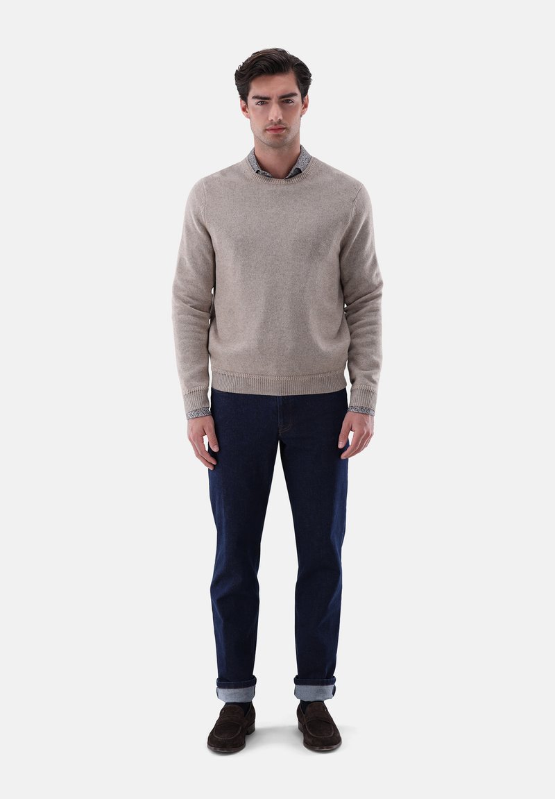 Beiger Strickpullover mit Rundhalsausschnitt, kombiniert mit dunkelblauen Jeans, hochgekrempelten Ärmeln und braunen Wildlederschuhen. Schlichtes Design, weicher Textil.