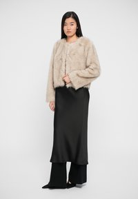 Beige faux fur lyhythihainen takki pitkillä hihoilla ja edessä napitus, käytetty mustan satiinisen liukupuvun päällä, yksinkertaista taustaa vasten.