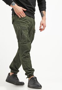 Cargo byxor i mörkgrön tyg med flera fickor, tajt modell vid anklarna och en avslappnad passform. Matchas med strukturerade svarta sneakers.