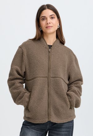 Femme portant une veste polaire marron avec fermeture éclair, les mains dans les poches, debout devant un fond clair uni.