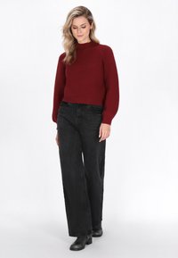 Czerwony sweter dzianinowy w prążki z wysokim kołnierzem, zestawiony z czarnymi spodniami z wysokim stanem i szerokimi nogawkami oraz czarnymi botkami za kostkę. Proste tło.