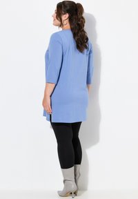 Hellblaues Tunika mit Dreiviertelärmeln, kombiniert mit schwarzen Leggings und grauen High-Heels-Stiefeln. Glatter Stoff mit seitlichen Schlitzen.