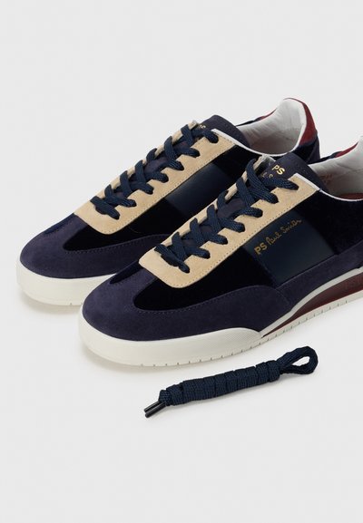 Sneakers in suede blu navy con dettagli beige, caratterizzati da un pannello lucido, lacci piatti blu navy e una suola in gomma bianca testurizzata. Include un paio di lacci extra.
