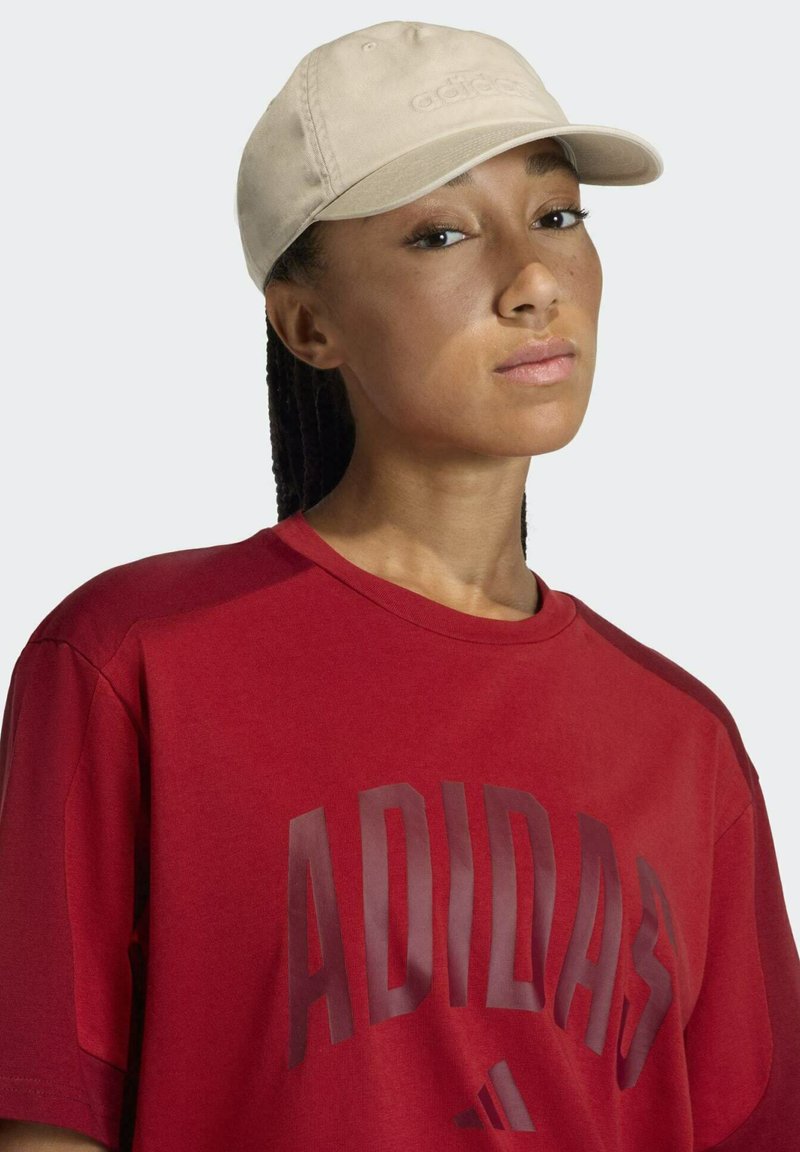 Gorra de algodón beige con visera curva y logo bordado, combinada con una camiseta de algodón roja que presenta un estampado oscuro y audaz.