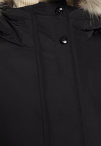 Svart vinterjacka närbild med glänsande svarta knappar och beige pälskant på huvens kant.