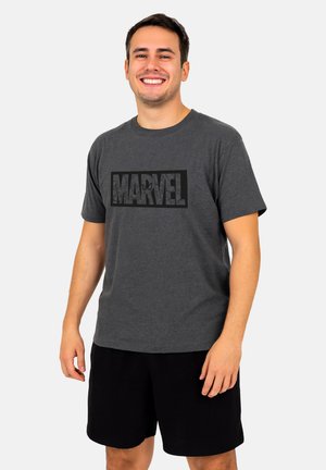 Uomo sorridente che indossa una maglietta nera con il logo Marvel e pantaloni corti neri, in piedi di fronte a uno sfondo chiaro e uniforme.
