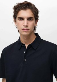 Čierna polokošeľa s textúrovanou tkaninou, golierom a tromi gombíkmi. Model má krátke vlnité vlasy a neutrálny výraz.