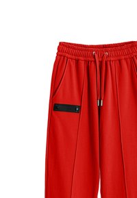 Pantalones jogger rojos con cintura elástica, cordón, bolsillos laterales y un acento de etiqueta negra en el bolsillo izquierdo. Textura de tela suave.