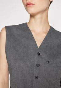 Szary kamizelka bez rękawów w jodełkę z dekoltem w kształcie litery V, z trzema czarnymi guzikami i małą kieszonką na piersi. Gładka tekstura.