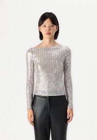 Ασημί μπλούζα με μακριά μανίκια φτιαγμένη από παγιέτες, με bateau λαιμόκοψη και εφαρμοστή σιλουέτα, συνδυασμένη με μαύρο δερμάτινο παντελόνι.