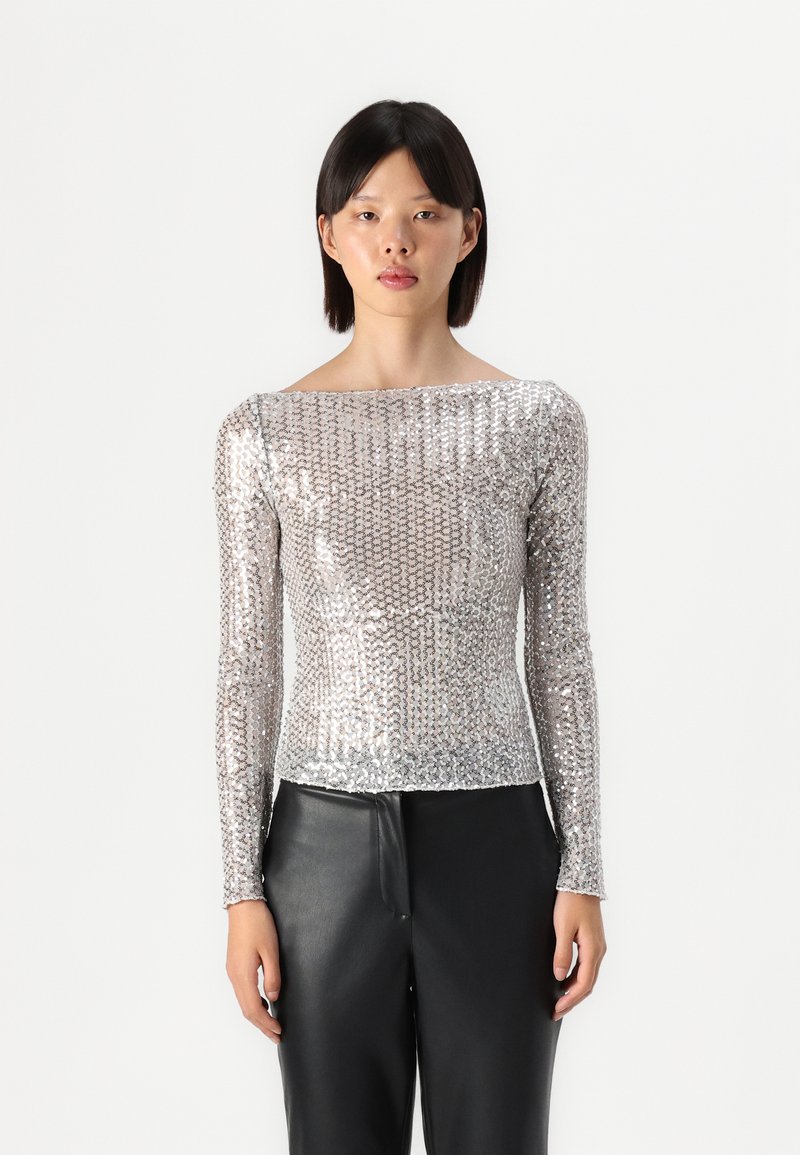 Ασημί μπλούζα με μακριά μανίκια φτιαγμένη από παγιέτες, με bateau λαιμόκοψη και εφαρμοστή σιλουέτα, συνδυασμένη με μαύρο δερμάτινο παντελόνι.