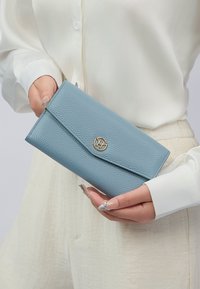 Portafoglio in pelle strutturata di colore azzurro chiaro con un accento in oro e chiusura a soffietto, tenuto in una mano, con una superficie liscia e bordi puliti.