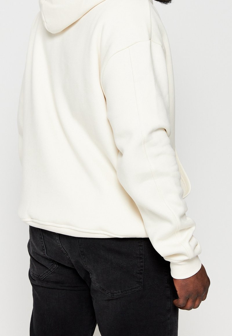 Sudadera con capucha de color crema con una textura suave, que cuenta con un bolsillo tipo canguro y puños acanalados, combinada con vaqueros de mezclilla oscura.